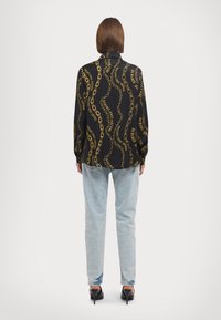 Blusa preta com corrente dourada e padrão de texto, mangas longas e tecido suave; combinada com jeans azul claro e saltos pretos.