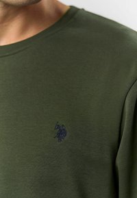 Tuva acs ar olīvu zaļas krāsas T-krekla audumu, uz kura krūtīm ir neliela tumša izšūta emblēma ar cilvēku, kas ceļ roku uz augšu.