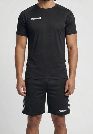 Homme portant un maillot de sport noir et un short avec des logos hummel blancs et des bandes chevron sur les côtés, debout devant un fond uni.