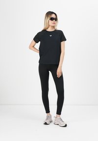Svart kortermet t-skjorte med et figursydd design og logo på brystet, kombinert med svarte leggings og lyse treningssko.