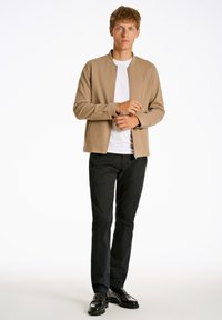Chaqueta cremallera en tono marrón clara, hecha de una tela suave sobre una camiseta blanca, combinada con pantalones negros y zapatos negros brillantes.