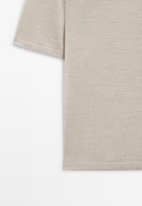 Tissu de chemise beige à manches courtes montrant une texture détaillée avec un ourlet cousu proprement sur fond blanc.
