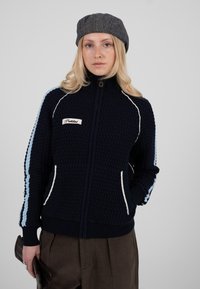 Veste en maille marine zippée avec motif texturé, rayures bleu clair et crème le long des manches, et poches avant. Détail de fermeture éclair ton or.
