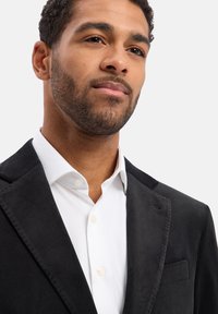 Blazer in velluto nero con rever a lancia e tasca frontale, abbinato a una camicia bianca con colletto e dettagli di bottoni visibili.