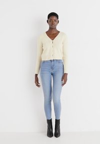 Lys beige strikket cardigan med V-hals og fire knapper, kombinert med lyseblå skinny jeans og svarte ankelstøvler.