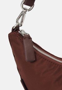 Bruine stoffen tas met een gladde textuur, zilveren ritssluiting en leren trekker, voorzien van een metalen gesp en riemaansluiting.