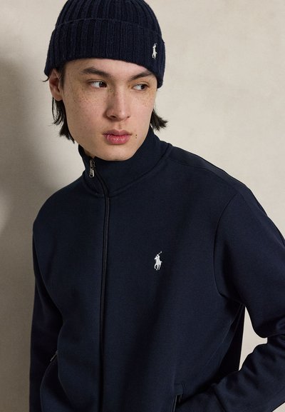 Young man with straight dark hair wearing a navy blue knit hat and matching zip-up jacket with a white polo logo, looking left.
