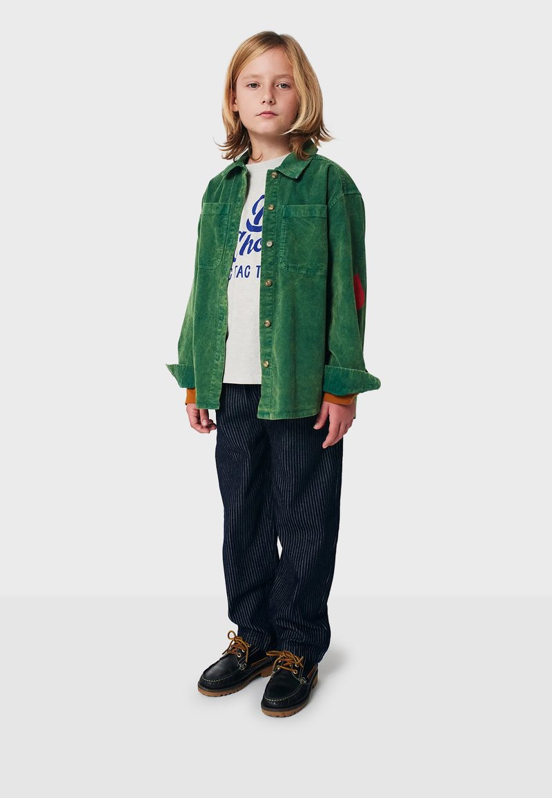 Grüne Cordhemd mit zwei Fronttaschen und einem roten Akzent; graues Grafik-T-Shirt darunter; gestreifte marineblaue Hose; schwarze Schnürschuhe.