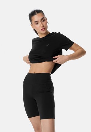 Nő felemeli a fekete rövid ujjú pólót, fekete magas derekú kerékpáros rövidnadrágot visel egyszerű fehér háttér előtt.