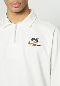 Camiseta polo blanca con collar y cremallera a medio camino, que presenta un logo bordado de Nike Sportswear en negro y naranja en el pecho. Textura de tela suave.