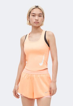 Ung kvinde med kort blondt hår iført en ferskenfarvet Nike tanktop og matchende træningsshorts, stående mod en ensfarvet baggrund.