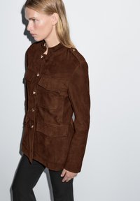 Veste en daim marron avec un col montant, une fermeture à boutons sur le devant et deux poches plaquées. Texture lisse et coupe ajustée.