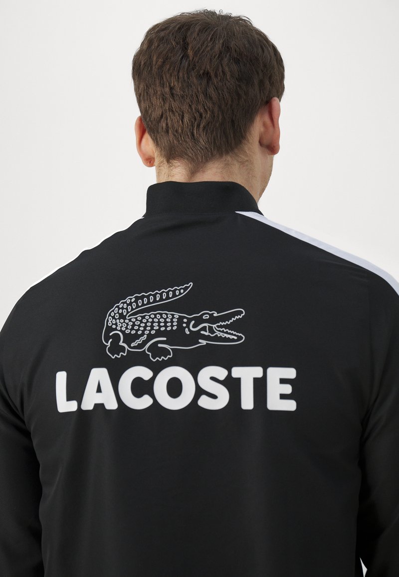 Giacca nera con colletto alto, caratterizzata da un logo Lacoste bianco e una grafica di coccodrillo sul retro. Materiale liscio con dettagli bianchi sulle maniche.