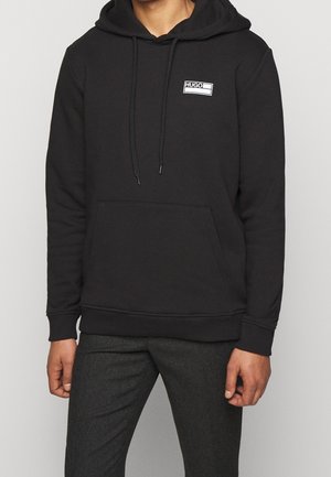 Man draagt een zwart Hugo hoodie met voorzak en aantrekkoord bij de capuchon, gecombineerd met donkere grijze broek.