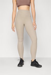 Leggings beige à côtes avec taille haute, associés à des chaussures de sport noires. Le tissu a une texture lisse et un design ajusté.