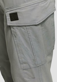 Lichtgrijze cargo shorts hebben een flapzak met een zwart reliëflogo. De stof is glad met zichtbare stikdetails en een relaxte pasvorm.