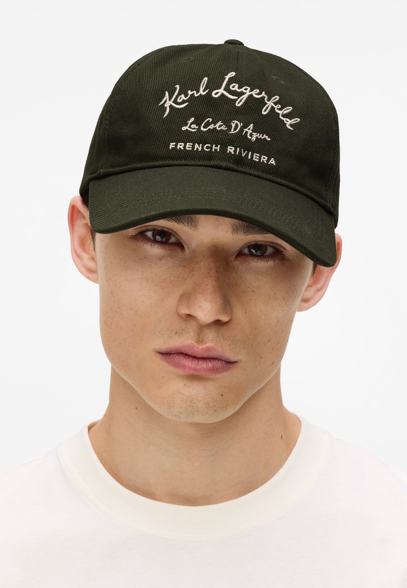 Casquette de baseball vert foncé avec texte blanc brodé indiquant "Karl Lagerfeld Le Côte D'Azur RIVIERA FRANÇAISE." Sangle réglable à l'arrière.