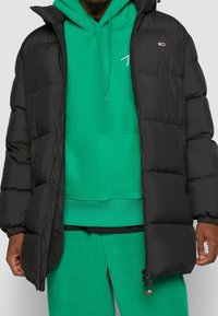 Svart puffjacka med huva, med dragkedja fram, elastiska muddars och framträdande sömmar, lager på lager över en grön hoodie.