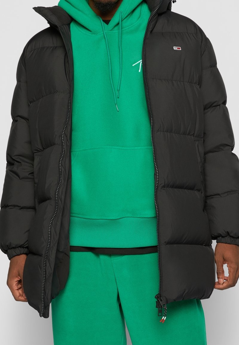 Svart puffjacka med huva, med dragkedja fram, elastiska muddars och framträdande sömmar, lager på lager över en grön hoodie.