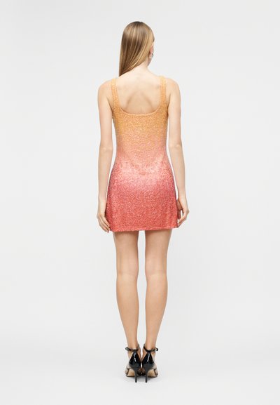 Γυναίκα που στέκεται γυρισμένη προς τα πίσω, φορώντας ένα μίνι φόρεμα χωρίς μανίκια με λαμπερή μετάβαση από πορτοκαλί σε ροζ και μαύρες ψηλοτάκουνες γόβες.