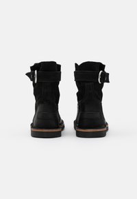 Botas de tobillo de ante negro con un puño doblado, acento de hebilla plateada y suela marrón contrastante. Presentan un acabado texturizado y un diseño minimalista.