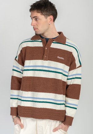 Férfi barna, fehér, zöld és kék csíkos pulóvert visel gallérral, balra néz, kezei a zsebében, semleges háttér.