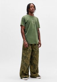 Camiseta verde oliva con un gráfico circular, combinada con pantalones verdes sueltos y estampados. Tejido texturizado, ajuste informal y zapatos de contraste.