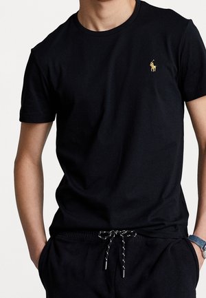 Mann trägt ein schlichtes schwarzes T-Shirt mit kurzen Ärmeln und kleinem goldfarbenem Polo-Spieler-Logo auf der Brust sowie schwarze Hosen mit Kordelzug, Hände in den Taschen.