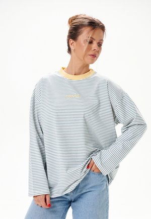 Femme avec les cheveux en chignon, portant une chemise rayée à manches longues avec un col jaune et un jean bleu clair, posant avec la main dans la poche sur fond blanc.