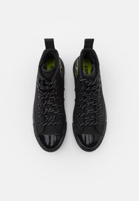 Zapatillas altas negras con punta de charol brillante, parte superior de tela texturizada, cordones en contraste y una plantilla verde brillante. Etiqueta negra en el talón.