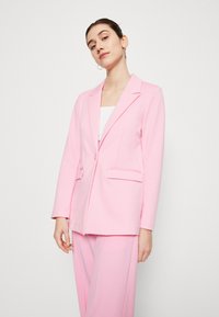 Blazer rose avec une fermeture à un bouton, des revers crantés, deux poches avant et une coupe ajustée, fabriqué à partir d'un tissu lisse.