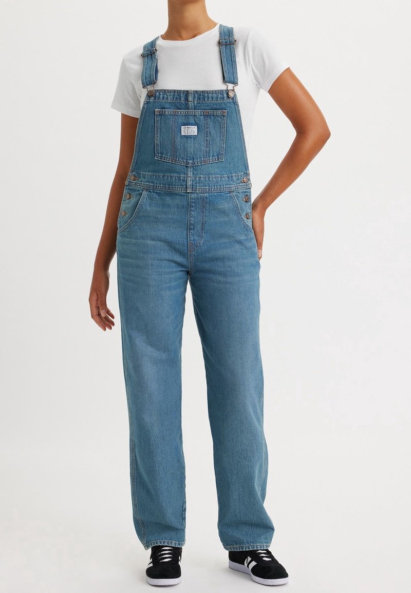 Salopette en denim bleu clair avec bretelles ajustables, poche avant, boutons sur les côtés et coupe ample, associée à un t-shirt blanc et des baskets noires.