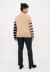 Woman facing away wearing a beige sweater with navy stripes on sleeves, black pants, and black heeled boots on a white background.