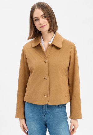 Femme aux cheveux bruns raides portant une veste marron claire boutonnée sur une chemise blanche et un jean bleu, sur un fond clair uni.