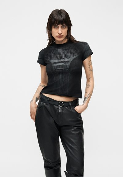 Άτομο που φοράει στενό μαύρο κοντομάνικο μπλουζάκι με σχεδιαστή υφή, ελαφρώς μισομπουκλωμένο σε μαύρο δερμάτινο παντελόνι ψηλόμεσο, με τα χέρια στις τσέπες.