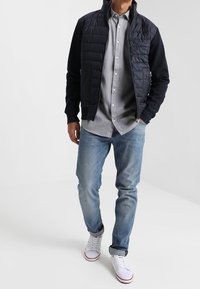 Chaqueta acolchada azul marino con cierre de cremallera sobre una camisa de botones gris claro, combinada con vaqueros azul claro y zapatillas blancas con detalles en rojo.