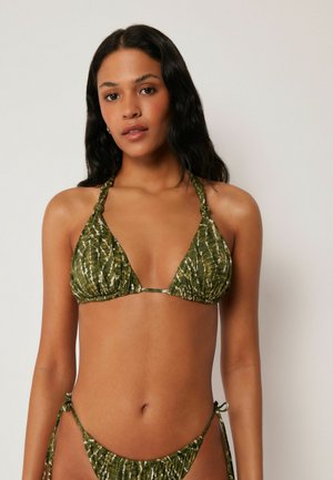 Kvinde iført grøn tie-dye halter bikini top med knyttede stropper og matchende bikini underdel bundet ved hofterne, stående mod en ensfarvet baggrund.