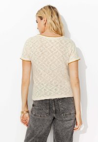 Haut en dentelle crème à manches courtes mettant en valeur un motif en diamant texturé et un contraste de bordures jaunes aux manches et au col. Porté avec un pantalon en denim gris.