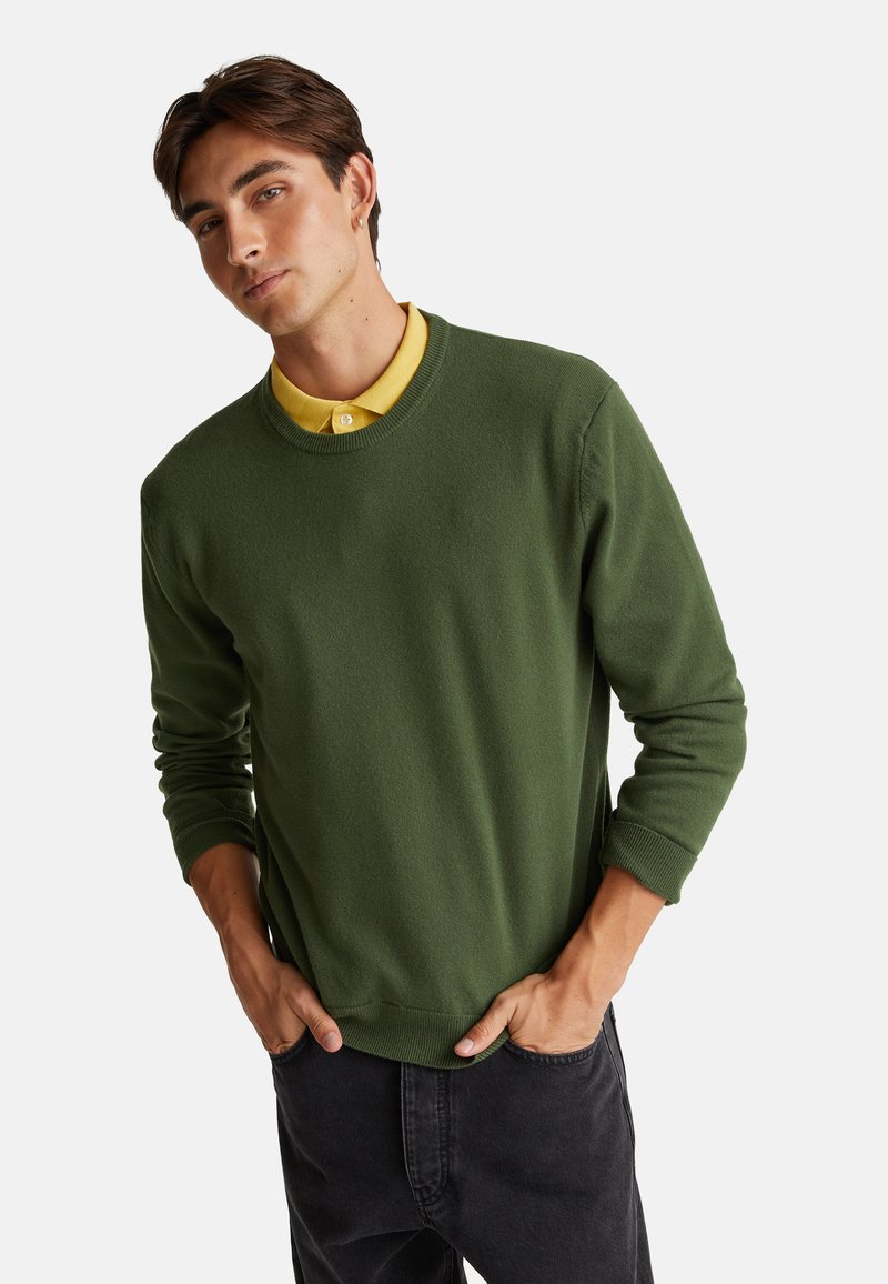 Giovane uomo che indossa un maglione verde a collo rotondo sopra una camicia gialla con colletto, con jeans neri, le mani in tasca, su uno sfondo chiaro e uniforme.