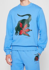 Persona que lleva un sudadera azul y pantalones con un gráfico de un cocodrilo verde y una flor roja, junto con los logotipos de Lacoste y Netflix.