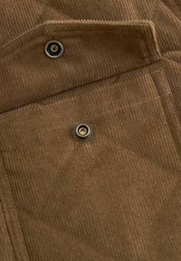 Gros plan sur un tissu en velours côtelé marron avec une poche à rabat sécurisée par deux boutons pression métalliques.