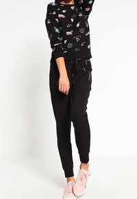 Sweatshirt noir avec des graphismes de dessins animés colorés, associé à un jogger noir et des baskets roses. La tenue présente une coupe décontractée et des poignets élastiques.