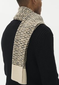 Strik scarf i beige og sort, med et tekstureret mønster. Enderne er ribbede i en ensfarvet kremfarve og hænger over en mørk sweater.