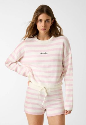 Femme portant un pull à rayures horizontales roses et blanches et un short assorti, debout avec une main sur la hanche devant un fond uni.