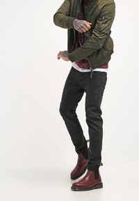 Chaqueta bomber verde oliva, camisa con patrón burdeos, jeans negros y botines Chelsea burdeos. Detalles notables incluyen un bolsillo con cremallera en la manga.
