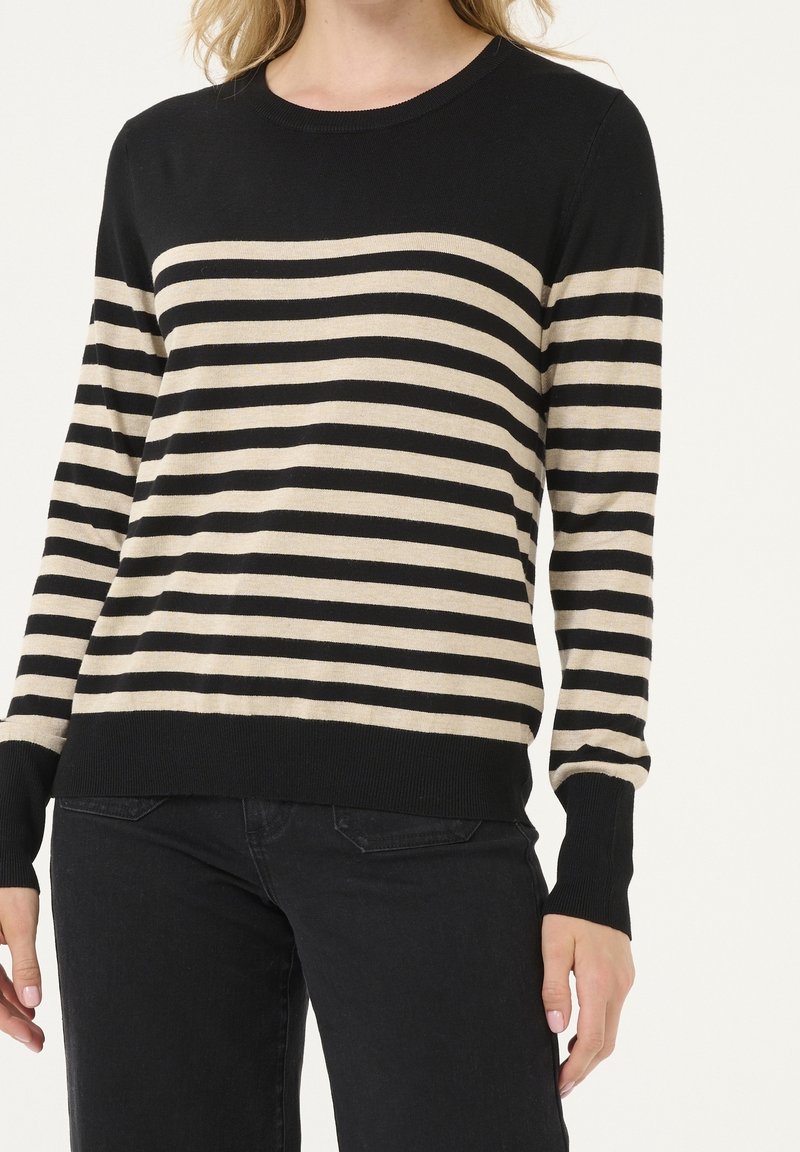 Femme portant un pull à manches longues rayé horizontal noir et beige et un pantalon noir, visible du cou aux hanches.