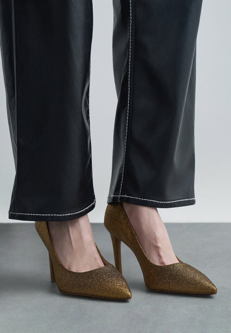 Zlatne šljokice visoke pete s oštrim prstima i stiletto petama, u kombinaciji s crnim hlačama širokih nogavica s bijelim šavovima.