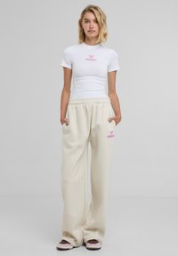 T-shirt bianca aderente con logo rosa, abbinata a pantaloni della tuta beige larghi con tasche laterali e logo rosa coordinato.