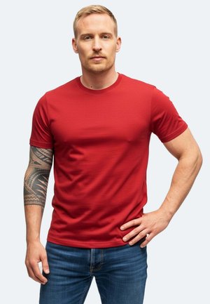 T-shirt rossa a maniche corte in tessuto liscio con scollo rotondo, abbinata a jeans in denim blu. Mostra un intricato tatuaggio sul braccio sinistro.