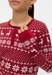 Kobieta w czerwonej bluzce z długim rękawem, z białymi wzorami reniferów i śnieżynkami, trzymająca kołnierz przy guzikach, nosząca perłowe kolczyki w kształcie kropli.